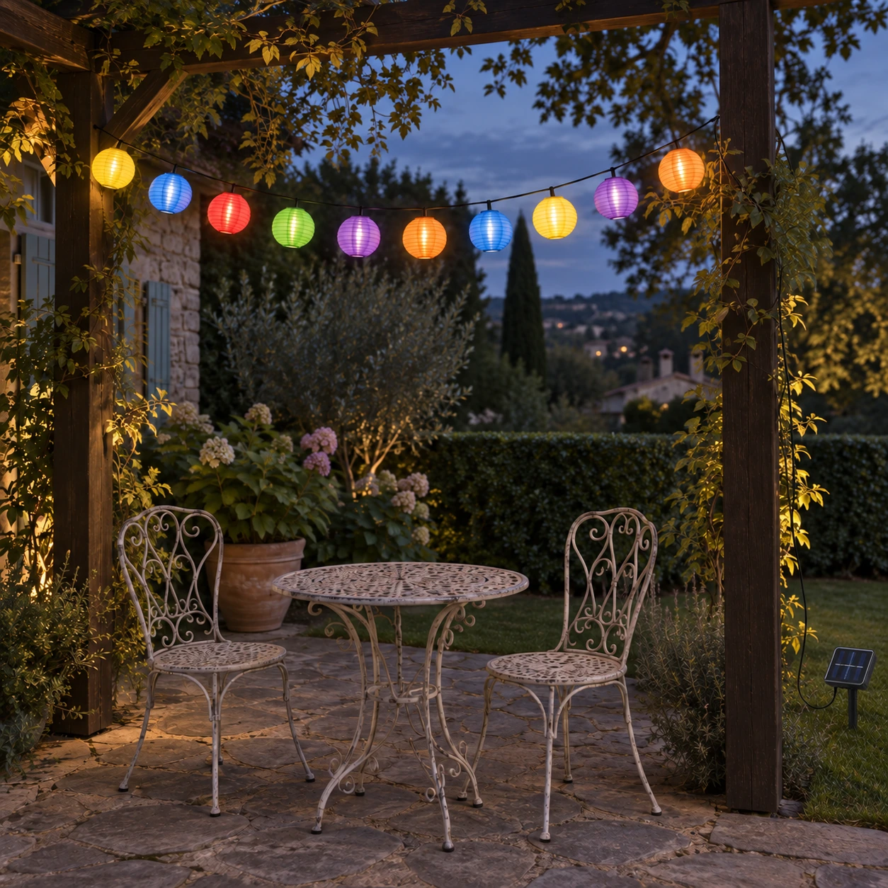 Dix lanternes colorées allumées sous une pergola, au-dessus d'une table de jardin en fer forgé