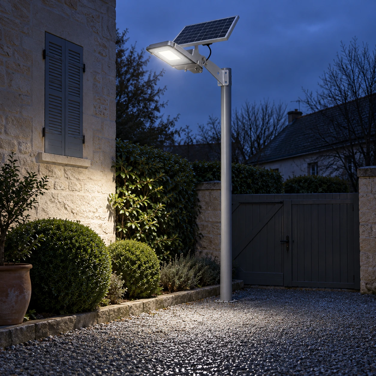 De nuit, le lampadaire solaire allumé sur son mât éclaire une cour en gravier devant une maison en pierre