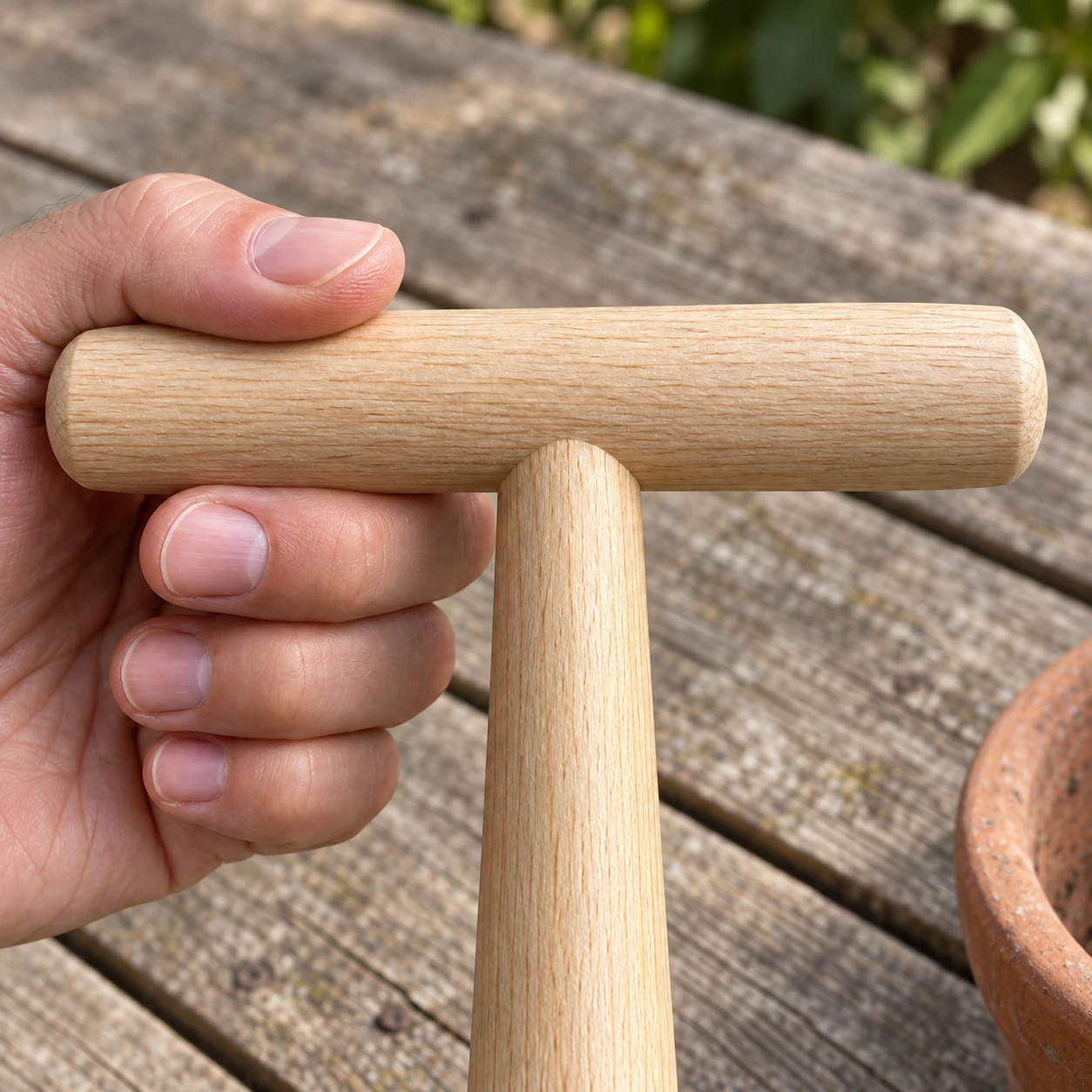 La traverse en bois clair du plantoir tenue dans une main, au-dessus d'une table de jardin